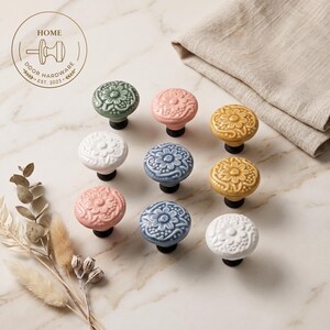 May include: A collection of nine round ceramic cabinet knobs arranged in a grid, featuring embossed floral patterns in sage green, soft pink, mustard yellow, white, and dusty blue, each mounted on a black base with a circular logo in the corner.