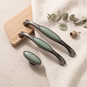 May include: Two metal drawer pulls and one matching cabinet knob, each featuring a dark gunmetal finish and a smooth, oval-shaped sage green inlay. The hardware pieces have a beaded border surrounding the central green oval accent.