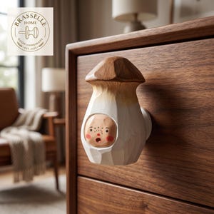 May include: A mushroom-shaped wooden drawer knob featuring a carved brown cap and a white stem. The stem includes a small, recessed face with painted eyes, freckles, and a red mouth. The knob is mounted on a dark wood drawer front.