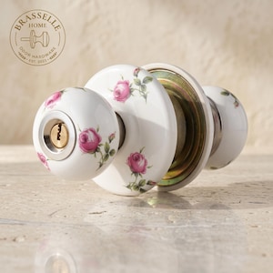 May include: A white porcelain door knob set featuring a delicate pink rose floral pattern with green leaves. The front knob includes a brass keyhole mechanism, while the circular backplate and secondary knob are also decorated with matching roses.