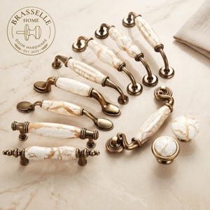 Peut inclure: Une collection de quincaillerie d'ameublement comprenant diverses poignées de tiroir et deux boutons ronds. Chaque pièce présente un centre en céramique blanche avec des veines de marbre doré et des accents en laiton antique. Logo Brasselle Home.