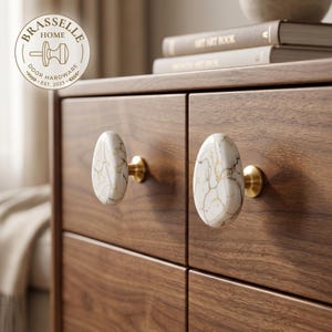 May include: Two oval-shaped cabinet knobs featuring a white marble-like surface with brown veining, mounted on a dark wood drawer unit. Each knob is attached with a circular brushed brass base. A circular logo reads BRASSELLE HOME DOOR HARDWARE.