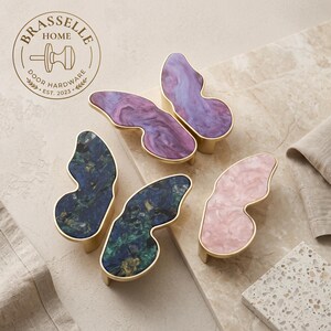Può includere: Quattro pomelli per armadi a forma di farfalla con bordi in metallo dorato e inserti in resina marmorizzata nei colori viola, blu scuro, verde e rosa chiaro. Il logo Brasselle Home Door Hardware Est. 2023 è visibile nell'angolo in alto a sinistra.