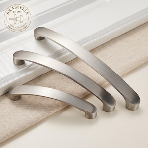 Pode incluir: Três puxadores de armário em níquel escovado de comprimentos variados dispostos na diagonal. Cada puxador apresenta um design arqueado suave com extremidades arredondadas e acabamento metálico. O logótipo Brasselle Home Door Hardware é visível.