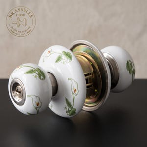 Peut inclure: Un ensemble de poignées de porte en céramique blanche orné d'un motif floral de lys calla peint avec des feuilles vertes et des pétales blancs. La quincaillerie comprend un insert de serrure métallique, une plaque de montage et un mécanisme interne.
