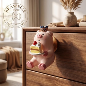 May include: A pink pig-shaped cabinet knob featuring a small blue and gold crown on its head and holding a white plate with a slice of cake topped with a red strawberry. The knob is mounted on a wooden drawer and includes a circular logo reading BRASSELLE HOME DOOR HARDWARE EST. 2023.