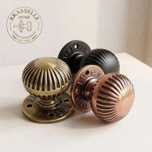 May include: Three round door knobs with a ribbed, fluted texture. One knob is finished in antique brass, one in matte black, and one in copper. Each knob features a circular mounting plate with visible screw holes and embossed lettering around the base.