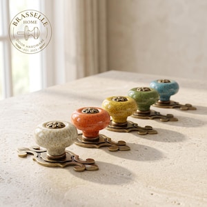 Può includere: Un set di quattro pomelli per porta in ceramica nei colori crema, arancione, giallo, verde e blu, con finitura maculata e un dettaglio floreale in metallo al centro. I pomelli sono montati su placche in ottone decorate in stile antico.