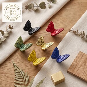 May include: Six butterfly-shaped cabinet knobs in black, red, green, gold, yellow, and blue. Each knob features detailed wing patterns. A logo in the top left corner reads BRASSELLE HOME DOOR HARDWARE EST. 2023 with a knob icon in the center.