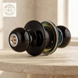 May include: A glossy black door knob set with a circular design and a central keyhole. The hardware features a metallic silver trim around the keyhole and a circular mounting plate with a brass-toned finish visible on the side of the mechanism.