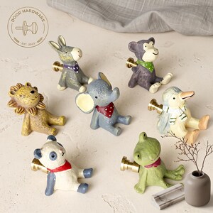 May include: A collection of seven animal-shaped drawer knobs featuring a lion, donkey, monkey, elephant, penguin, panda, and frog. Each knob is crafted with a brass-colored metal base and painted in distinct colors, including a red, green, or purple scarf.