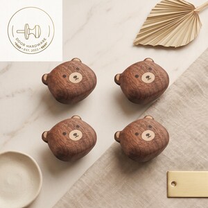 May include: Four dark brown wooden cabinet knobs shaped like bear heads with light wood accents on the face. Each knob features a small bear nose, mouth, and eyes. A circular logo in the top left corner displays a knob icon and the text Door Hardware Est. 2023.