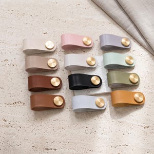 Puede incluir: Una colección de doce tiradores de cuero para cajones en varios colores, incluyendo blanco, rosa, lavanda, beige, gris, verde menta, marrón, negro, verde oliva, azul claro y amarillo mostaza, cada uno fijado con un cierre de latón cepillado.
