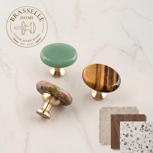 May include: Three cabinet knobs with polished brass bases and smooth, oval-shaped stone tops in green, brown tiger's eye, and mottled pink and green. The Brasselle Home Door Hardware logo is visible in the top left corner, and three material swatches are shown.