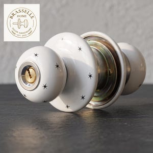 May include: A white ceramic door knob set featuring a black starburst pattern. The front knob includes a brass-toned keyhole cylinder. The hardware is branded with a circular logo in the corner that reads BRASSELLE HOME DOOR HARDWARE EST. 2023.