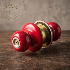 May include: A bright red door knob set featuring a spherical handle with a central keyhole, a circular brass-toned backplate, and a matching red knob on the opposite side. The hardware includes a silver-toned trim around the keyhole mechanism.