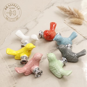 Puede incluir: Un conjunto de siete pomos para armarios de cerámica con forma de pájaro en blanco, amarillo, rosa, naranja, azul claro, verde claro y gris. Cada pomo tiene un acabado brillante, texturas de alas detalladas y una base de metal plateado.