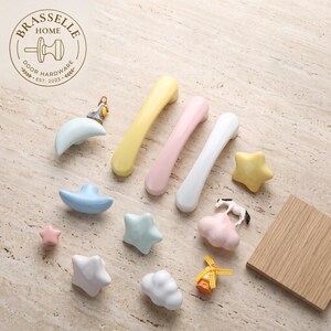 May include: A collection of pastel-colored cabinet hardware including three elongated handles in yellow, pink, and white, alongside various knobs shaped like stars, clouds, and moons, some featuring small decorative figures of a person, horse, and windmill.