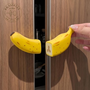 May include: Two yellow banana-shaped cabinet knobs with brown spots and a flat end showing a cross-section of the fruit. The left knob is attached to a wood-grain cabinet door, while the right knob is held against the adjacent door. Logo reads BRASSELLE HOME DOOR HARDWARE EST. 2023.