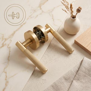 May include: A brushed gold door handle set featuring two cylindrical levers with a textured knurled grip pattern. The hardware includes circular mounting plates and an exposed internal mechanism. A line art icon of a door knob is visible in the corner.