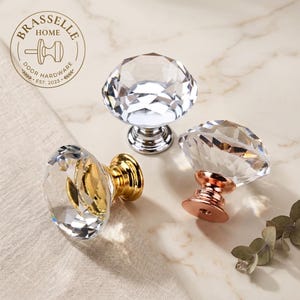 May include: Three faceted clear glass cabinet knobs with metallic bases in silver, gold, and rose gold finishes. Each knob features a multi-faceted, diamond-like top. A circular logo with the text BRASSELLE HOME DOOR HARDWARE EST. 2023 is visible.