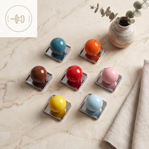 May include: A set of seven cabinet knobs, each featuring a smooth, spherical top in a distinct solid color including teal, orange, brown, red, pink, yellow, and light blue, mounted on a square, reflective silver-toned metal base plate.