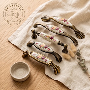 May include: Five cabinet pulls with antique brass-toned metal ends and central white ceramic inserts featuring floral patterns of yellow and red roses. The logo BRASSELLE HOME DOOR HARDWARE EST. 2023 is visible in the upper left corner of the image.