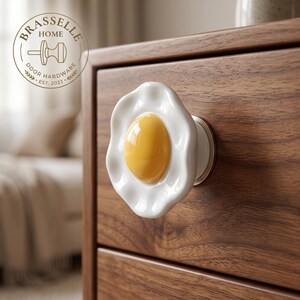 May include: A ceramic cabinet knob shaped like a fried egg with a glossy white scalloped edge and a raised yellow center, mounted on a dark wood drawer. The logo BRASSELLE HOME DOOR HARDWARE EST. 2023 is visible in the upper left corner.