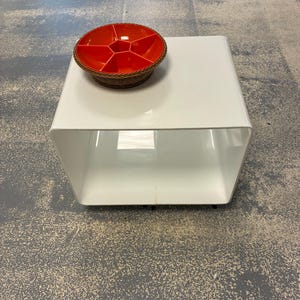 Puede incluir: Una mesa auxiliar cuadrada y hueca de color blanco con acabado brillante. Sobre ella descansa un plato de servicio redondo, naranja, con múltiples compartimentos y un borde de mimbre trenzado marrón. La mesa tiene una estructura abierta.