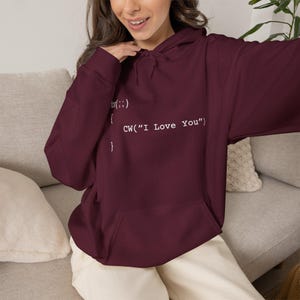 Peut inclure: Un sweat-shirt à capuche bordeaux profond avec une impression graphique blanche d'un extrait de code de boucle de programmation qui indique for(;;) { CW("I Love You") }. Le vêtement comprend une poche kangourou, des manches longues.