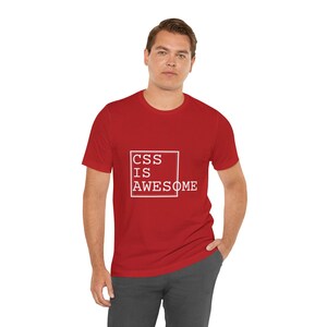 May include: A red short-sleeved t-shirt featuring a white square outline on the chest. Inside the square, the text reads CSS IS AWESOME in a white, block-style font. The shirt has a crew neckline and a standard fit, providing a simple design.