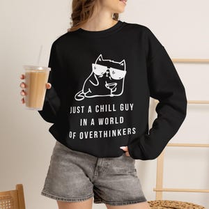 May include: A black long-sleeve sweatshirt featuring a white graphic of a cat wearing sunglasses and holding a drink. Below the cat, the text reads: JUST A CHILL GUY IN A WORLD OF OVERTHINKERS. The sweatshirt is paired with gray denim shorts.