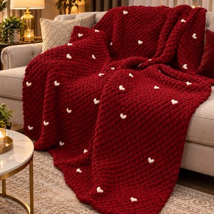 Puede incluir: Una manta de punto grueso en color rojo oscuro con un patrón repetitivo de pequeños corazones blancos en relieve. La manta presenta un tejido texturizado y voluminoso, extendida sobre un sofá, mostrando su construcción de hilo suave.