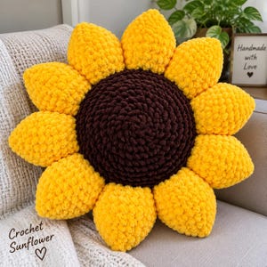 Puede incluir: Un cojín redondo y afelpado con forma de girasol, hecho de lana gruesa amarilla y marrón oscuro. El centro es un círculo texturizado marrón oscuro rodeado de segmentos amarillos brillantes en forma de pétalos. Se ve el texto "Crochet Sunflower".