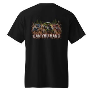 Op de afbeelding: Een zwart t-shirt met korte mouwen en een afbeelding op de achterkant van drie terreinwagens in blauw, groen en rood die door modder rijden met dennenbomen op de achtergrond. De tekst CAN YOU HANG staat in het wit onder de voertuigen.