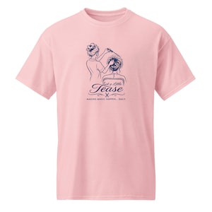 Puede incluir: Una camiseta de color rosa claro de manga corta con un gráfico azul oscuro de una persona peinando a otra. El texto dice Just a Little Tease y MAKING MAGIC HAPPEN... DAILY. El diseño incluye unas pequeñas tijeras bajo la palabra Tease.