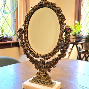 May include: An oval tabletop mirror featuring an ornate metallic gold frame with sculpted cherub and floral details. The mirror is supported by a central pedestal base that rests on a rectangular white marble plinth, providing a stable foundation.