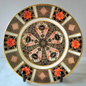 May include: A round ceramic plate featuring a central floral motif with black and orange petals on a dark blue background. The border displays alternating panels of orange flowers and geometric grid patterns, all accented with gold trim.