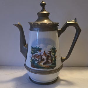 May include: A white ceramic coffee pot featuring a colorful landscape illustration of a castle and trees. The pot has a metallic lid, handle, and base, with a scalloped edge around the lid. The design includes a tall spout and a finial on top.