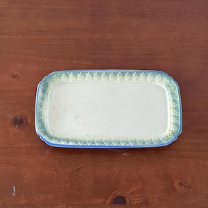 May include: A rectangular ceramic tray with a smooth, cream-colored center and a raised, textured border featuring a repeating leaf pattern. The rim is finished with a gradient of soft green and blue tones, creating a distinct edge.