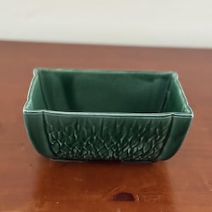 May include: A rectangular ceramic bowl with a deep forest green glaze. The exterior features a raised, textured pattern resembling woven leaves or scales, while the interior is smooth with a glossy finish and slightly flared edges.