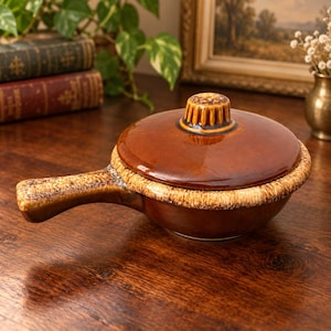 May include: A brown ceramic pot with a matching lid and a single handle. The pot features a smooth, glossy dark brown finish on the lid and center, contrasted by a textured, speckled beige and brown rim around the edge of the vessel.