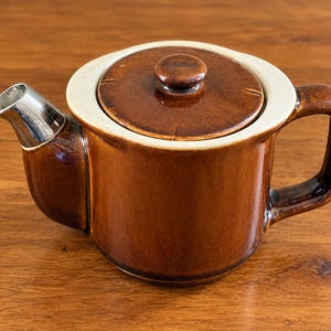 May include: A brown ceramic teapot featuring a glossy finish, a matching lid with a small knob, a sturdy side handle, and a metallic silver-toned spout. The teapot has a wide, cream-colored rim encircling the top opening and the lid.