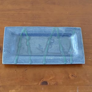 May include: A rectangular ceramic tray with a mottled blue glaze finish. The surface features two vertical, wavy lines in a lighter teal green color that run from the top edge to the bottom edge of the tray, creating a distinct contrast.