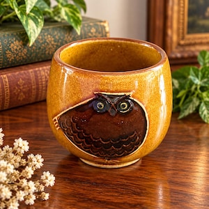 May include: A round ceramic mug with a glossy mustard yellow glaze and a dark brown embossed owl design on the front. The owl features detailed feathers and two prominent eyes. The mug has a smooth, curved shape with a wide opening and a solid base.