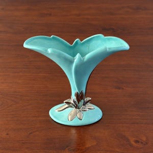 May include: A turquoise ceramic vase with a wide, flared, petal-like rim and a narrow neck. The base of the vase features a decorative silver-colored metallic accent shaped like a cluster of leaves or petals, resting on a circular pedestal.
