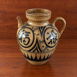 May include: A tan ceramic pitcher with a speckled finish, featuring a handle, a small spout, and black hand-painted circular motifs containing Chinese characters. The base is decorated with a black triangular pattern around the circumference.
