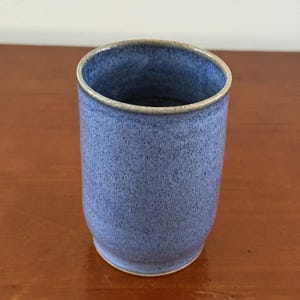 May include: A cylindrical ceramic tumbler featuring a speckled blue glaze with a smooth, light-colored rim. The vessel has a slightly tapered base and a consistent, mottled texture across its exterior surface, showcasing a handcrafted appearance.