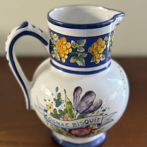 May include: A white ceramic pitcher featuring a blue rim and handle. The upper section displays a yellow grape and green leaf pattern. The body shows a floral design with a blue ribbon banner containing the text COGNAC BISQUIT.