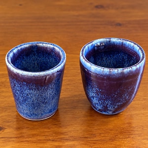 May include: Two ceramic cups featuring a reactive glaze with deep blue and purple tones. The finish displays a mottled, speckled texture with lighter blue highlights around the rims and upper sections of the cylindrical vessels.